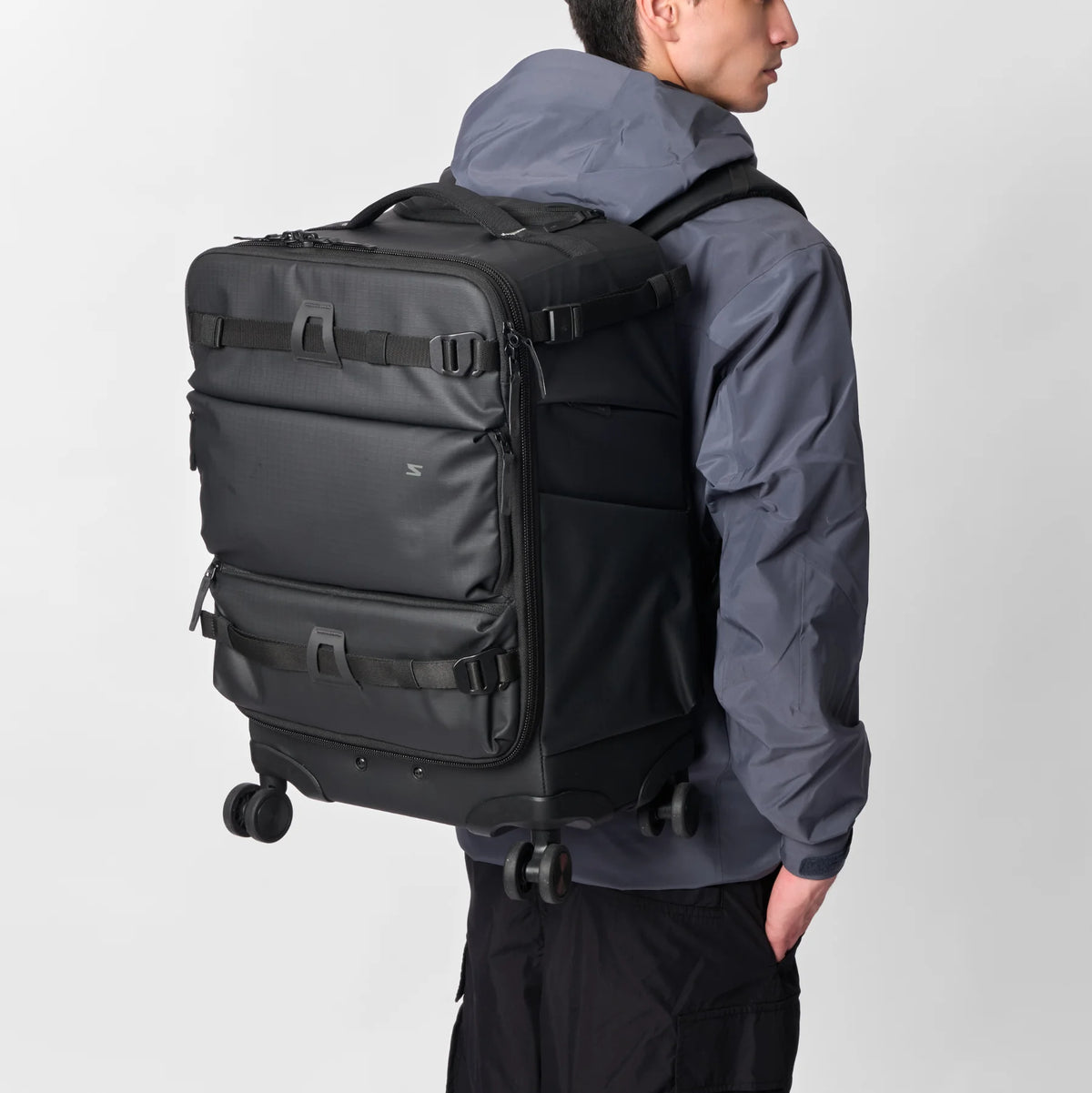 A man wearing a hooded jacket and black pants, carrying a large black rolling suitcase backpack with multiple compression straps and wheels against a light gray studio background.