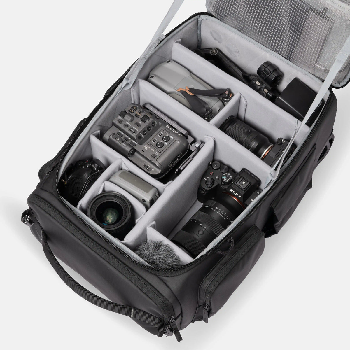 An open black camera gear bag with gray foam compartments, shown from above on a light gray background. The bag is filled with cameras, lenses, a mixer, and other photography accessories.