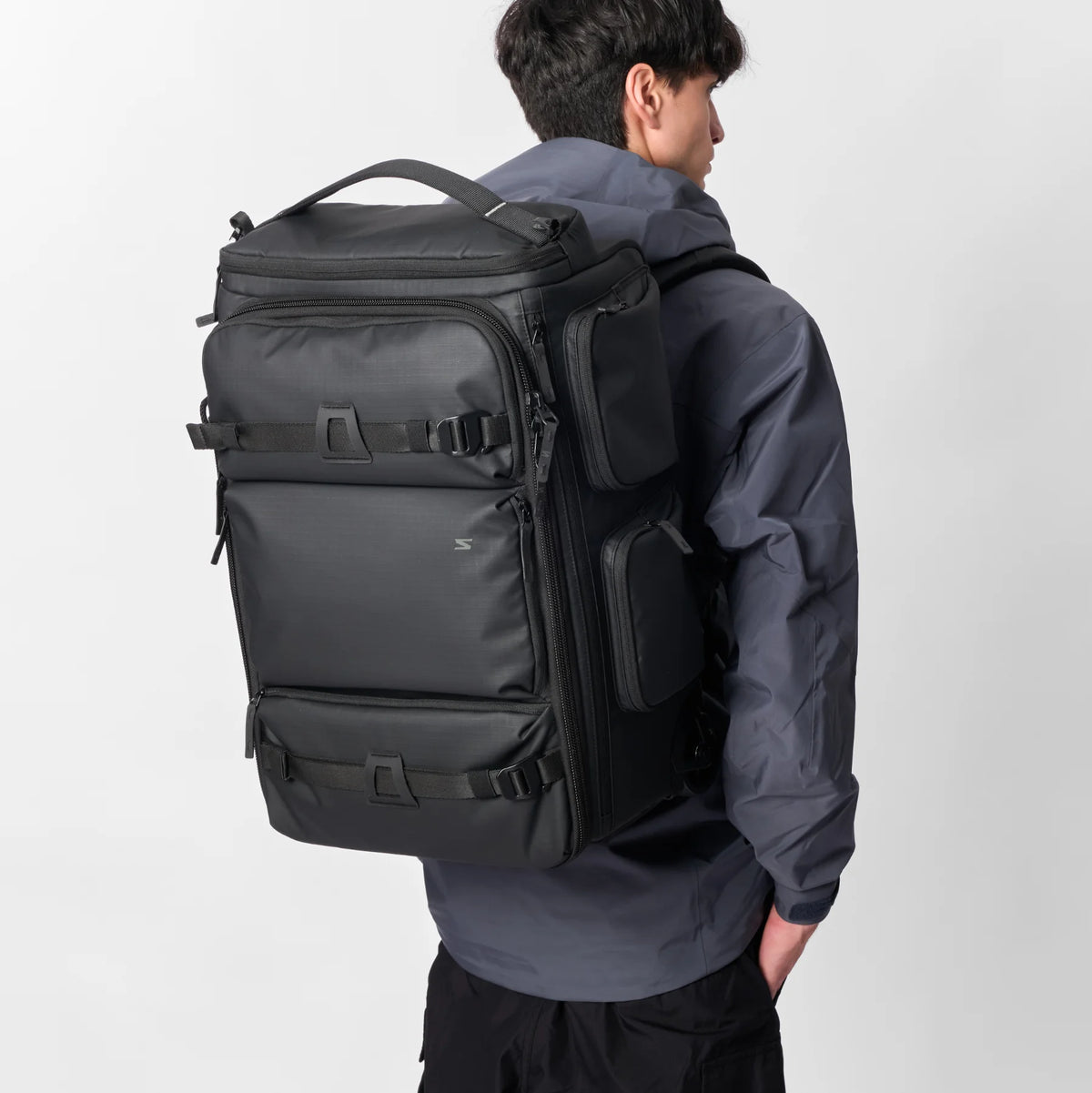 A man wearing a dark gray hooded jacket and black pants, shown from the back while wearing a large black travel backpack with multiple zippered compartments and compression straps.