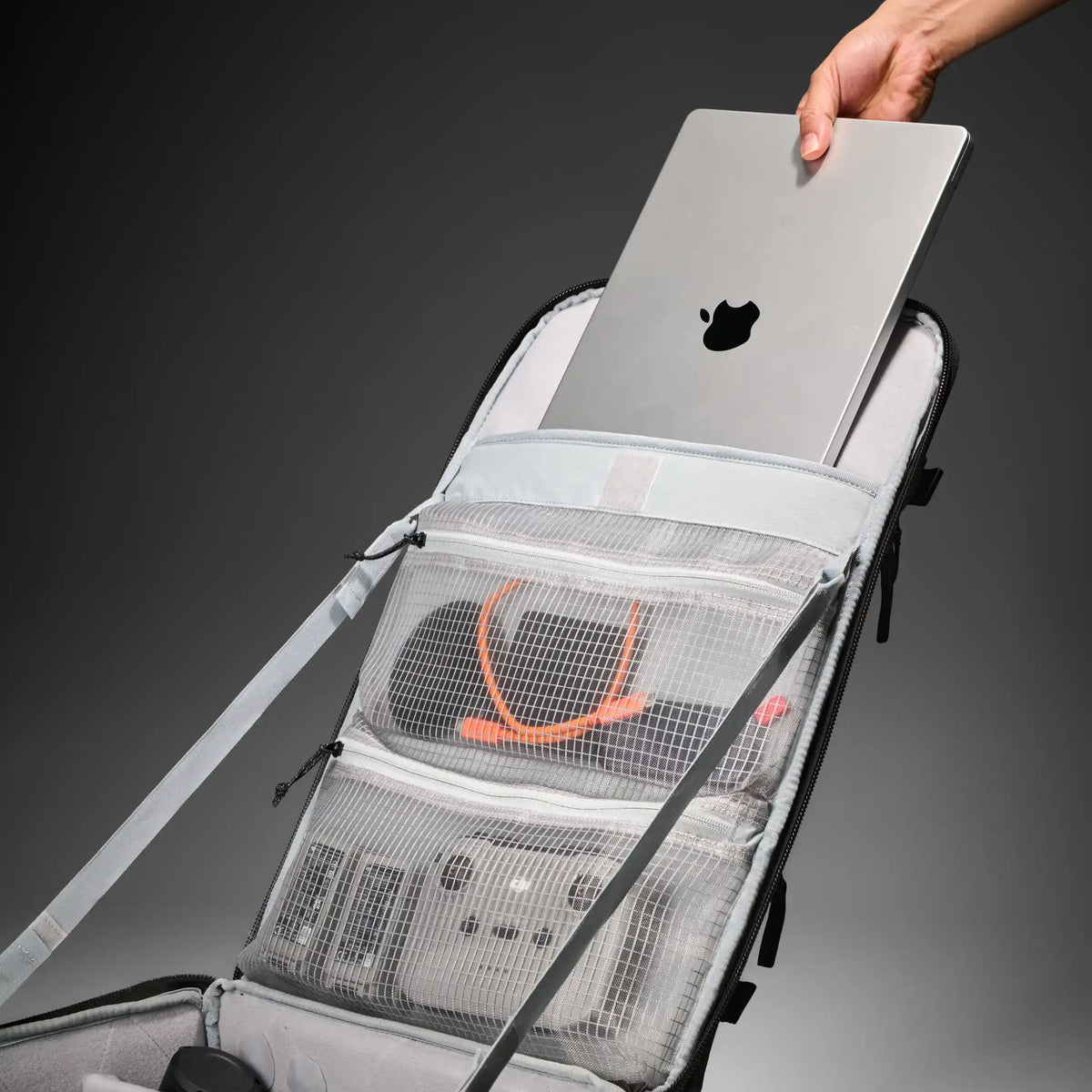 An open gray and black camera backpack with mesh pockets containing camera gear, with a hand holding a silver laptop inside. The background is a clean dark gray studio gradient.