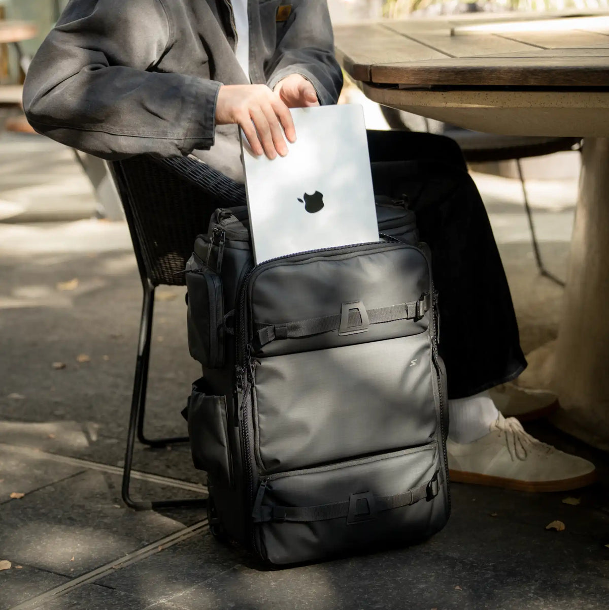 A person sitting at an outdoor table holds a silver laptop into the open top of a large black backpack-style bag. The bag has multiple zip compartments and front straps, shown in a real patio setting.