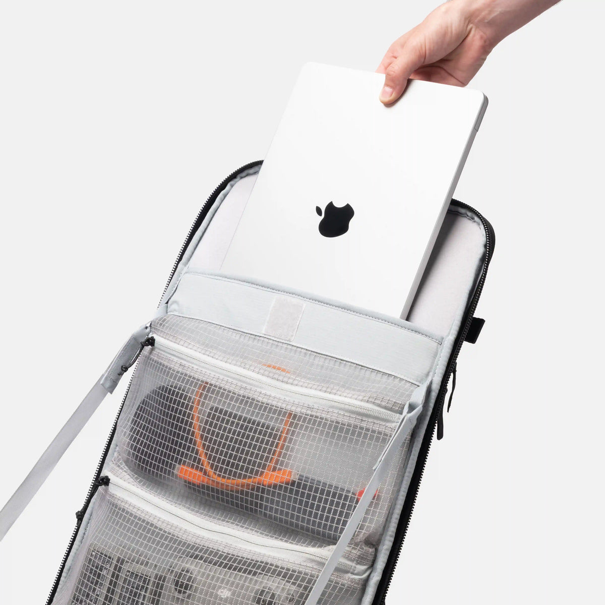 A light gray laptop backpack with a hand inserting a white laptop into its top compartment. The backpack features a clear mesh organizer pocket with orange accents and zippered compartments, shown against a solid light gray background.