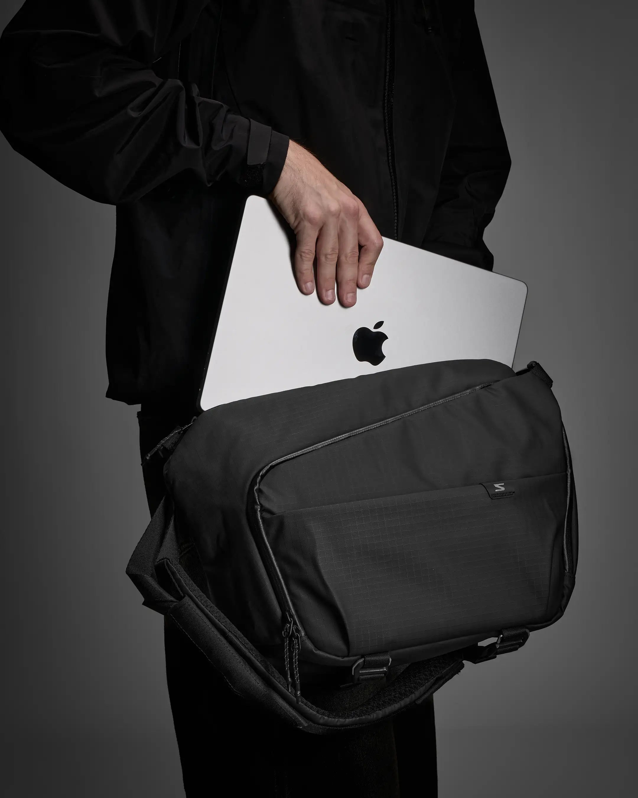 Person holding a black bag with an Apple laptop inside on a gray background
