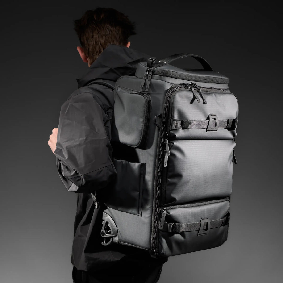 A man wearing a black hooded jacket is shown from the back, carrying a large black rolling duffel-style backpack with multiple zip compartments and straps against a dark gray studio background.