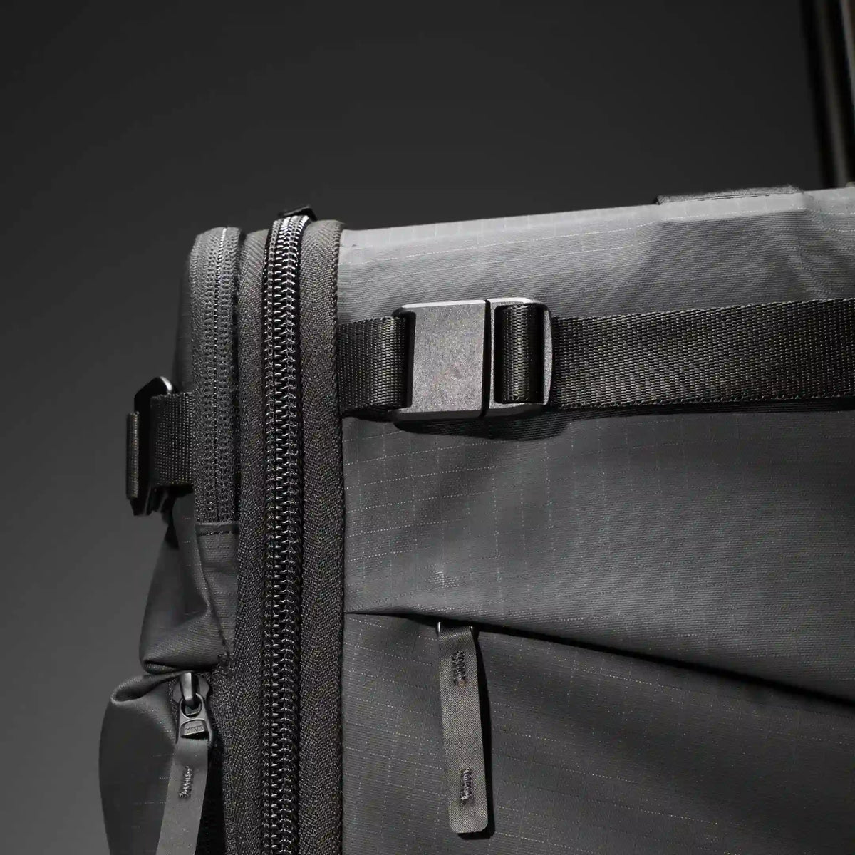 Close-up of a dark gray rolling suitcase with a telescoping handle extended, showing a front zipper, compression straps with black plastic buckles, and a small silver logo near the bottom.