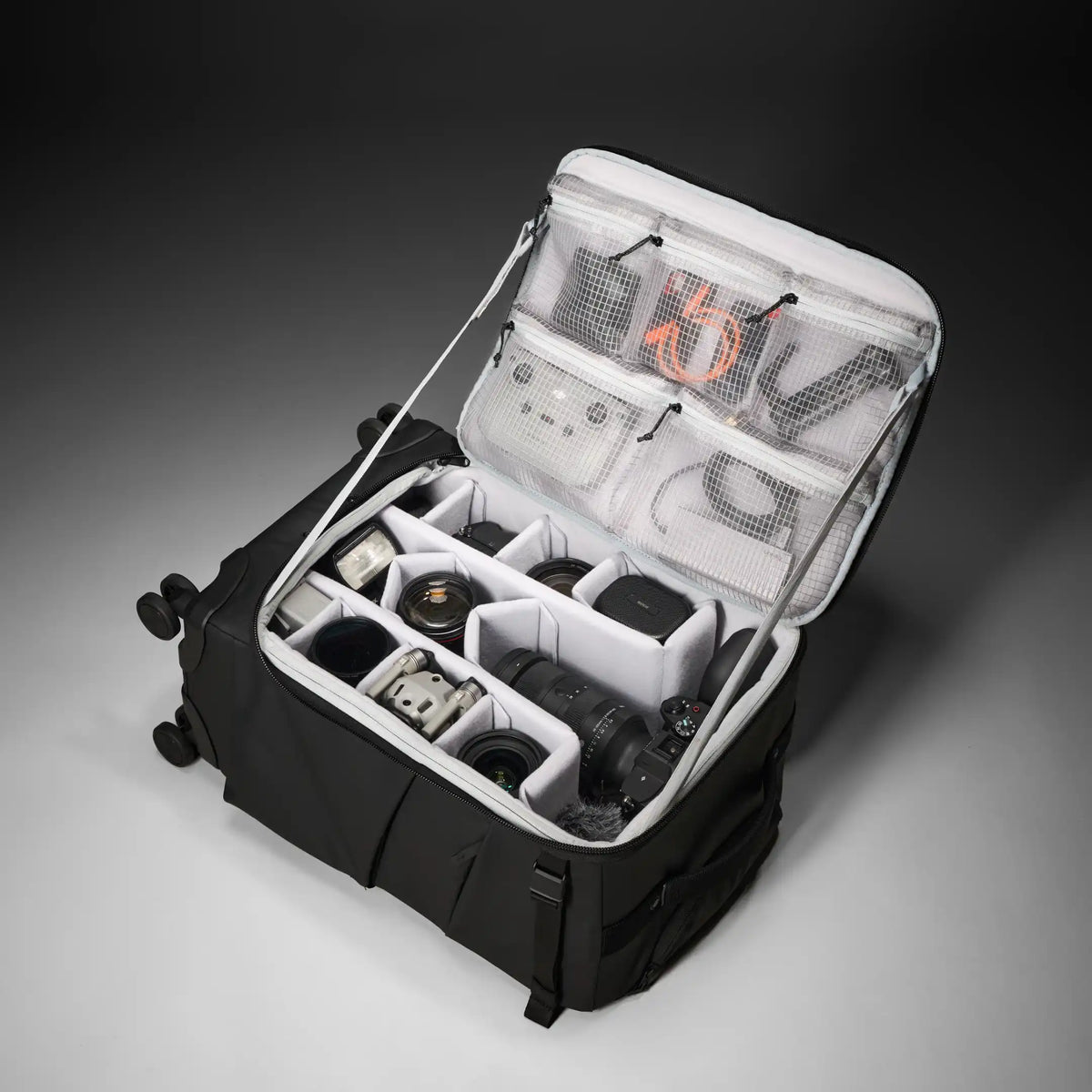 An open black camera gear organizer bag with gray interior compartments, shown holding camera lenses, a camera body, and accessories. The bag is photographed on a dark studio background.