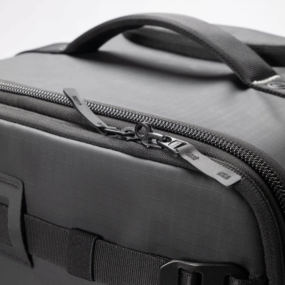 Close-up of a dark gray textured soft-sided bag with a top handle, black zipper, and silver zipper pull on a light gray background.