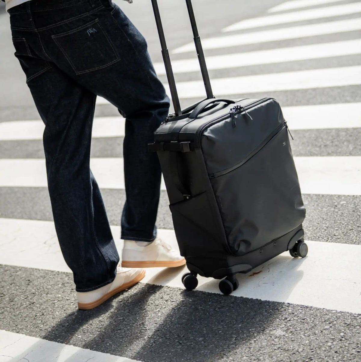 A person in dark blue jeans and white sneakers pushes a black rolling suitcase with an extended telescoping handle across a zebra crosswalk.