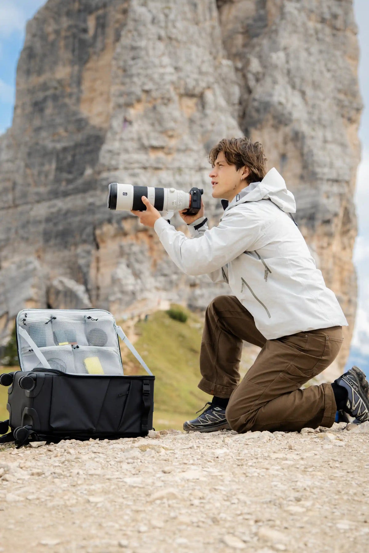 A man in a white jacket and brown pants kneels on a rocky mountain path holding a camera with a long telephoto lens, with an open black gear bag beside him and a large rock formation in the background.
