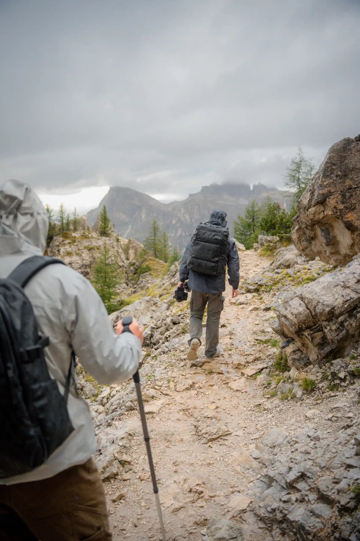 Two hikers on a rocky mountain trail under a cloudy sky, with rugged peaks in the background. One person in the foreground wears a light gray hooded jacket and black backpack, using a trekking pole, while another hiker walks ahead in the distance.