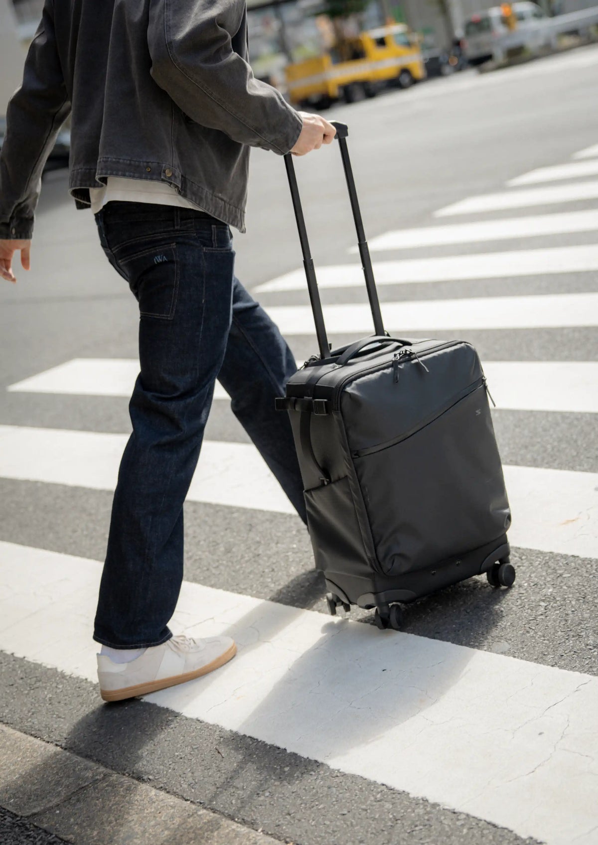A person walking across a crosswalk pulling a dark gray rolling suitcase with an extended handle.