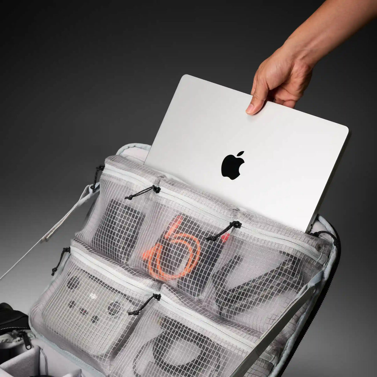 A gray mesh laptop organizer bag with multiple zippered compartments and pockets, shown holding a silver Apple MacBook and other gear. A hand is visible sliding the laptop into the top compartment.