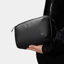 Person holding a black crossbody bag with a brand logo on a light gray background