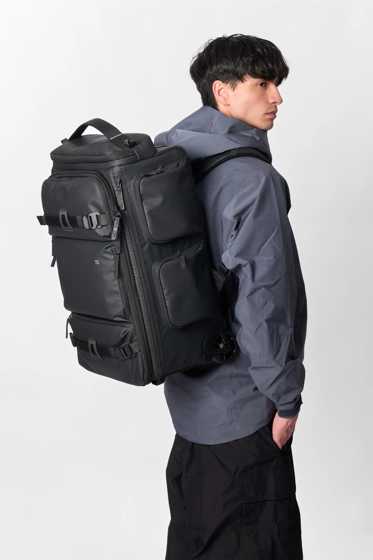 A man wearing a dark gray hooded jacket and black pants, shown from the back and side, carrying a large black backpack with multiple compartments and straps against a light gray studio background.