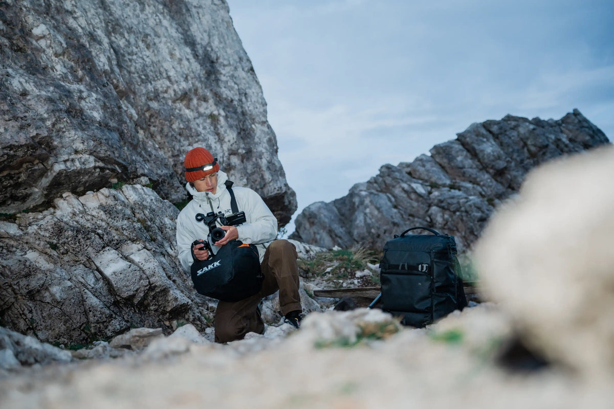 A man in outdoor gear, including a white jacket, brown pants, and an orange beanie, sits on a rocky mountain ledge holding a black bag. A larger black backpack is placed on the rock beside him, with a blurred foreground rock in the foreground.