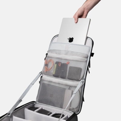 A gray and black camera gear backpack shown open, with a hand inserting a silver laptop into the top compartment. The backpack has multiple mesh and padded organizer pockets containing camera accessories.