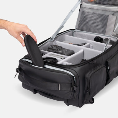 A black camera gear organizer bag with a gray interior organizer system, shown open with a hand placing a black case inside. The bag features multiple compartments, a side pocket, and a mesh lid panel, photographed on a clean light gray background.