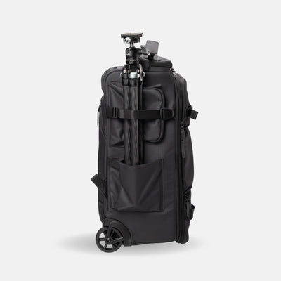 A black rolling camera bag with a tripod attached to its side, shown from a three-quarter rear view against a light gray studio background.