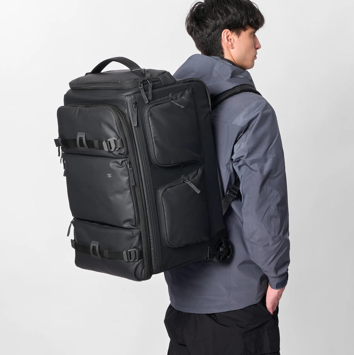 A man wearing a dark gray jacket and black pants is shown from the back wearing a large black rolling travel backpack with multiple zippered compartments and side straps, set against a plain light gray studio background.