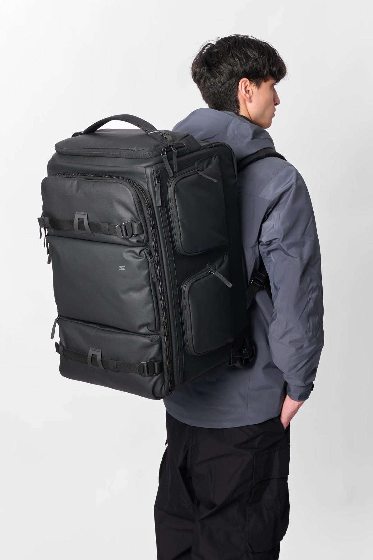 A man wearing a dark gray jacket and black pants is shown from the back, carrying a large black rolling travel backpack on his shoulders against a light gray studio background.