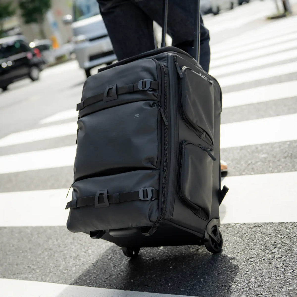 A black rolling suitcase with multiple zippered compartments and buckle straps, being pulled by a person on a city crosswalk.