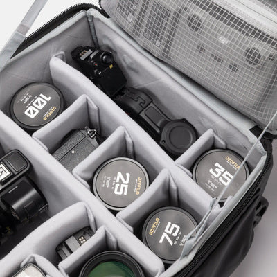 An open black camera gear bag with gray foam compartments, shown holding camera lens caps and camera accessories. The bag's interior organizer is clearly visible, along with several lens caps featuring gold and white markings.