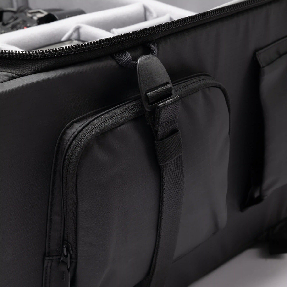 Close-up of a black soft-sided travel or equipment bag with a front zippered pocket and an adjustable hanging strap, shown against a light neutral background.