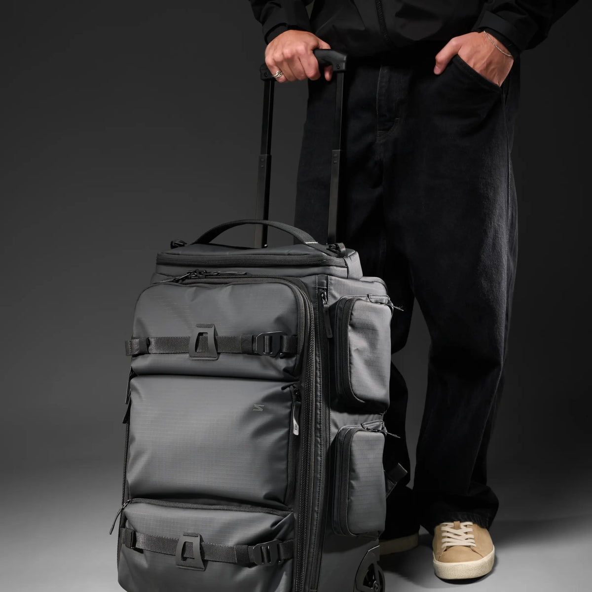 A dark gray rolling duffel bag with multiple zippered compartments and side pockets, shown next to a person wearing black pants and beige sneakers against a dark gray studio background.
