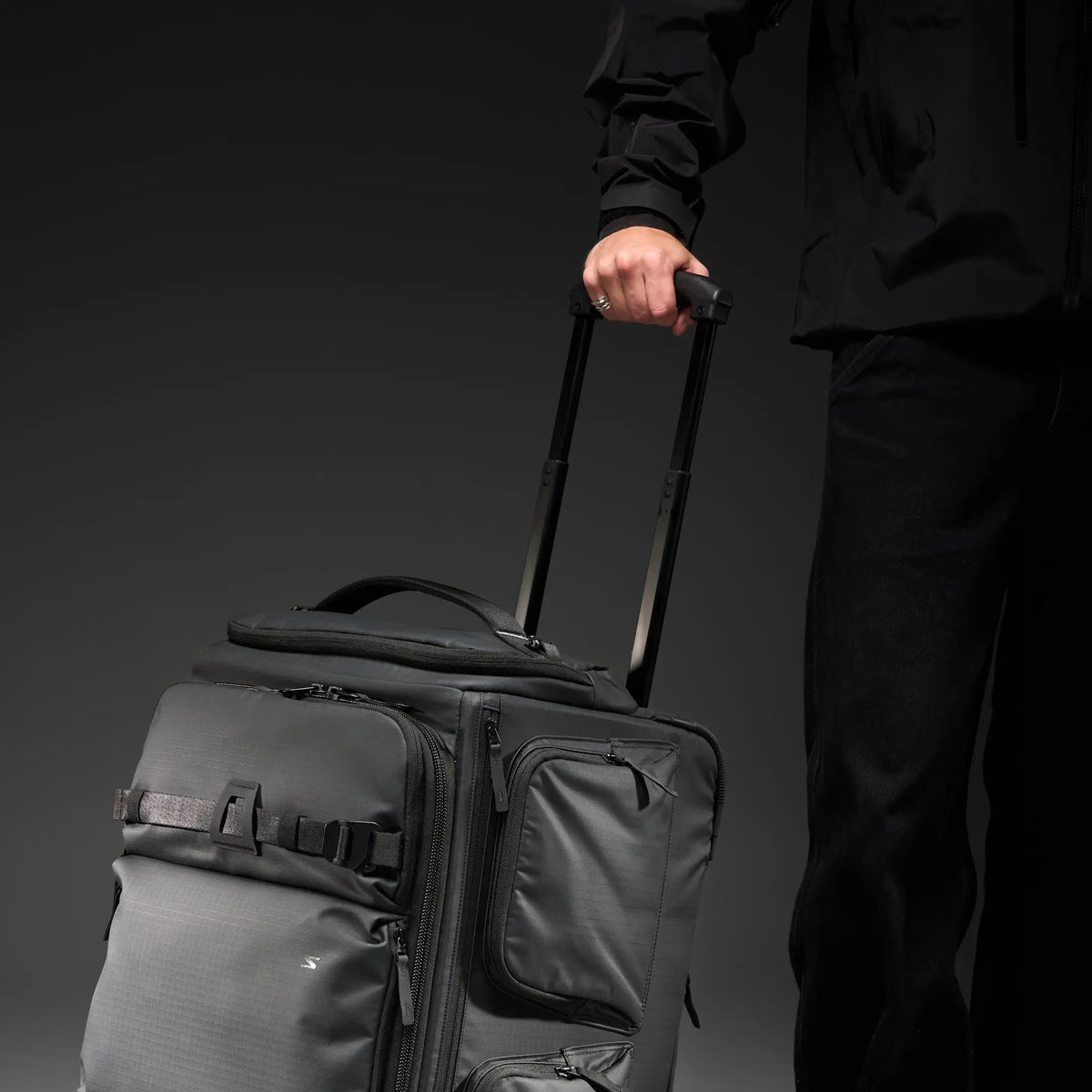 Close-up of a dark gray soft-sided rolling suitcase showing the textured fabric, a recessed telescoping handle, and part of a front zipper pocket.