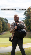 Man with glasses and beard wearing a plaid shirt using a Sakk Gear bag to support his video camera with a fuzzy microphone outdoors; text overlay says "saving my back with the SAKK"
