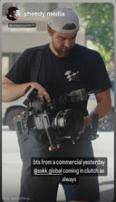 Instagram story screenshot showing a man in a backwards cap operating a large camera rig with a Sakk Gear bag, captioned "bts from a commercial yesterday @sakk.global coming in clutch as always"