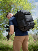 Man wearing a large black Sakk Gear backpack outdoors, holding a professional camera rig in his hand among green trees