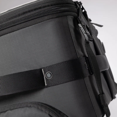 Close-up of a black technical backpack showing the textured ripstop fabric, a black webbing strap with a snap button closure, and a zipper compartment.