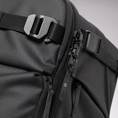 Close-up of a black textured backpack showing a side zipper, black plastic zipper pulls, a black webbing strap with a metal buckle, and a black zipper tab.