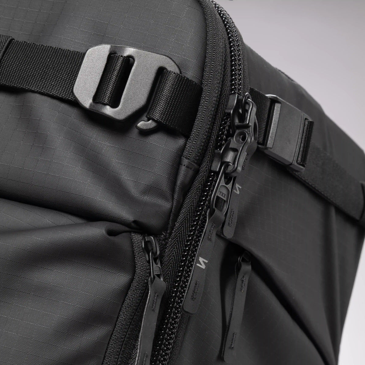 Close-up of a black textured backpack showing a side zipper, black plastic zipper pulls, a black webbing strap with a metal buckle, and a black zipper tab.
