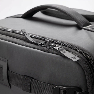 Close-up of a dark gray textured soft-sided bag with a top handle, black zipper, and silver zipper pull on a light gray background.