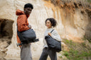Two people carrying black duffel bags in a natural setting with rocky cliffs.
