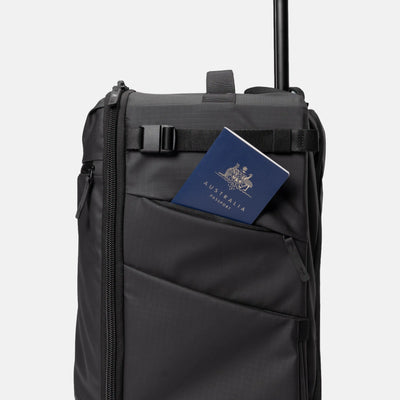 A black rolling suitcase with an extended telescoping handle, featuring a front utility pocket holding a blue passport under a black strap, set against a plain light gray background.