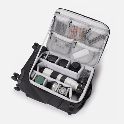 An open black and gray camera gear organizer suitcase shown from a top-down angle, filled with camera equipment including a telephoto lens, camera body, and accessories in mesh and foam compartments.