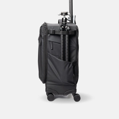 A black soft-sided travel bag with wheels and an extended telescoping handle, shown from the back with a tripod mounted on the handle wrap, against a light gray studio background.