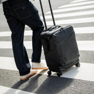 A person in dark blue jeans and white sneakers pushes a black rolling suitcase with an extended telescoping handle across a zebra crosswalk.
