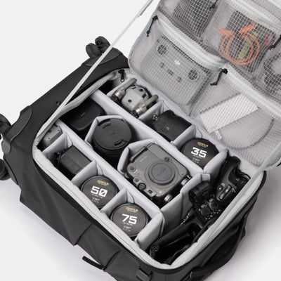 An open black camera gear bag with gray interior organizer compartments, shown on a white studio background. Visible items include lens caps labeled Sigma 35mm, 50mm, and 75mm, a camera body, a lens, and mesh pockets with accessories.