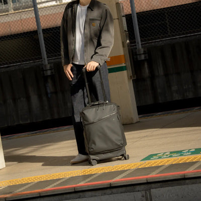A person standing on a train platform holding the handle of a dark olive green rolling suitcase. The person is wearing a dark gray zip-up jacket over a white t-shirt and dark jeans.