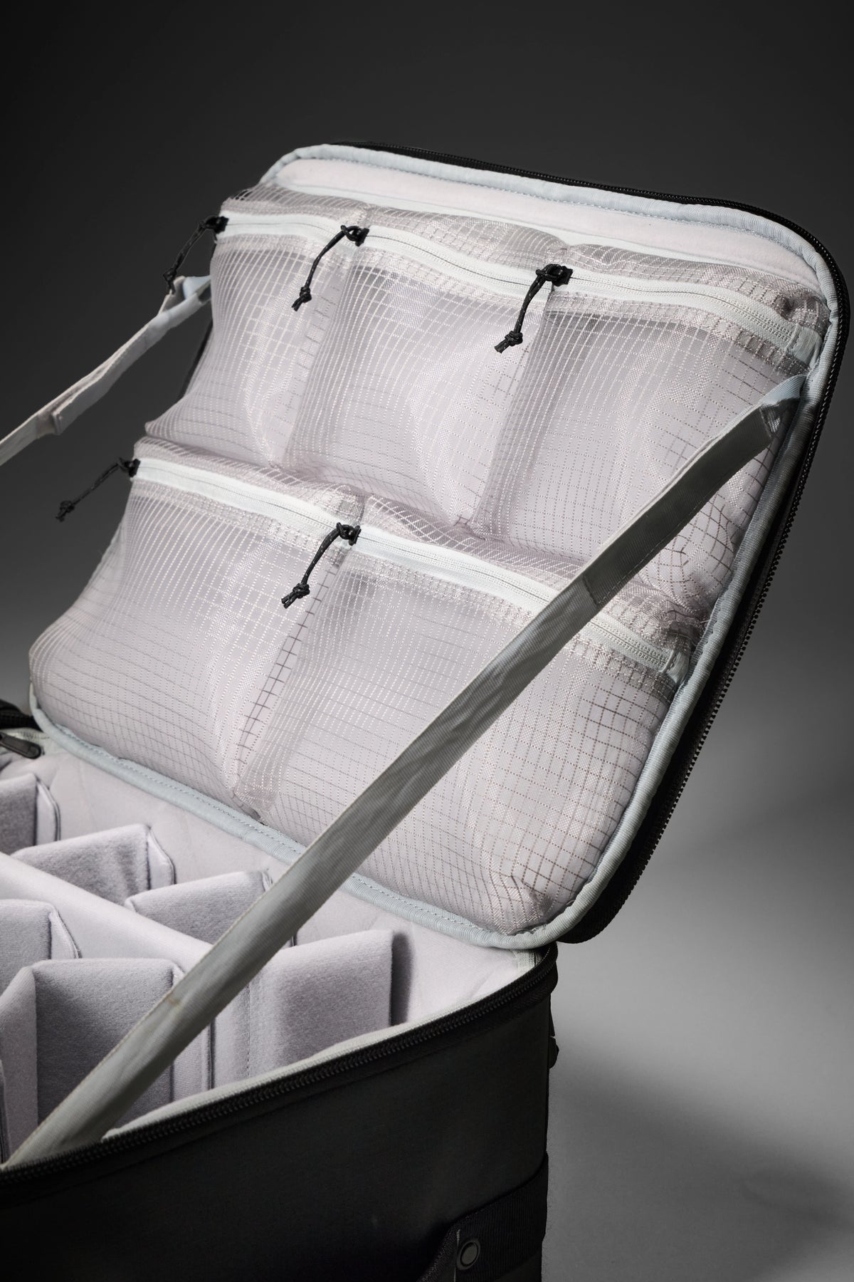 An open black and gray luggage organizer suitcase showing white mesh zippered compartments and padded dividers inside, photographed on a dark gray studio background.