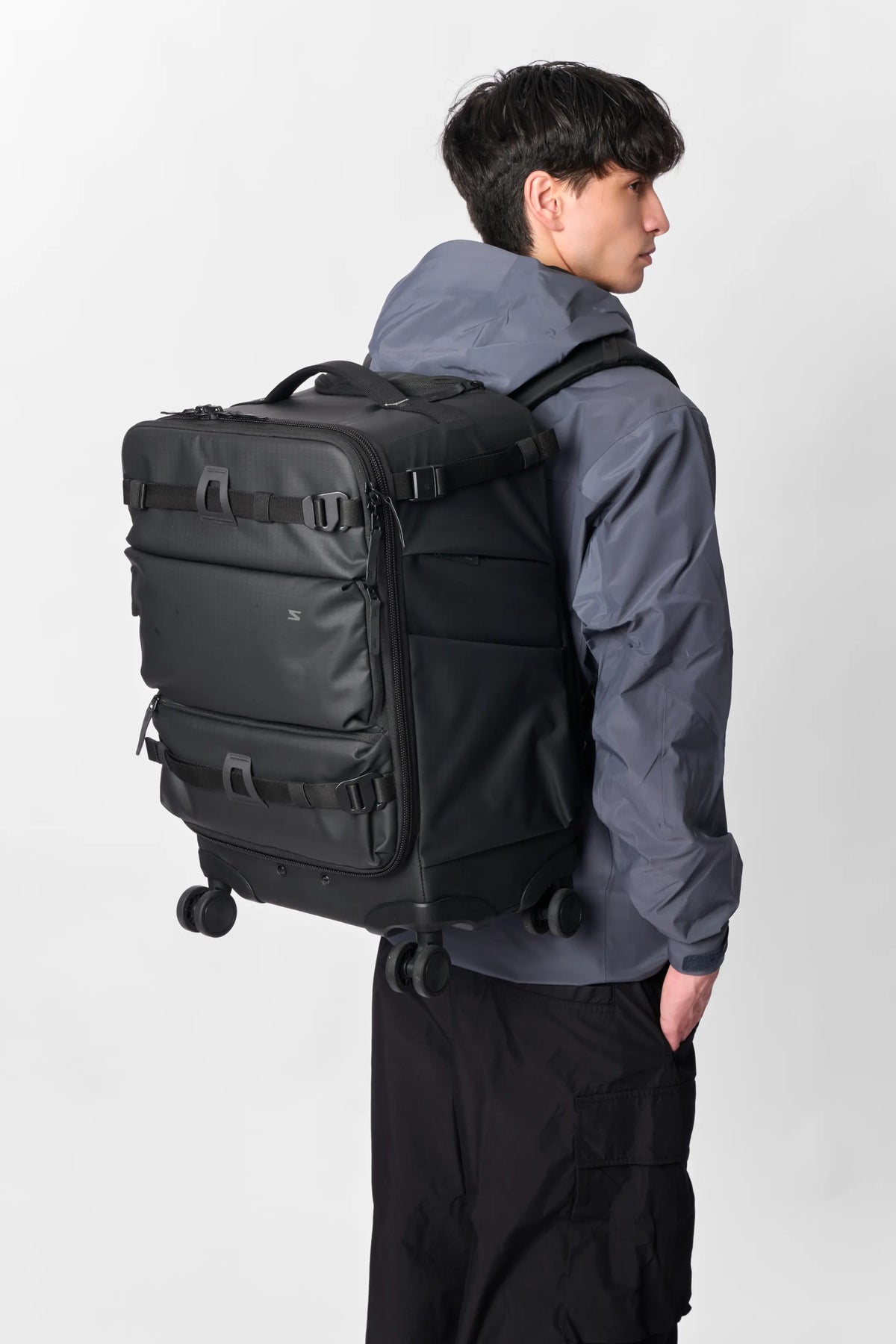 A man wearing a dark gray hooded jacket and black cargo pants, shown from the back and side, wearing a black rolling duffel backpack with multiple compression straps and wheels.