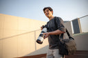 Outdoor photographer using Nomad sling bag while holding DSLR with telephoto lens. Secure compartments for gear.