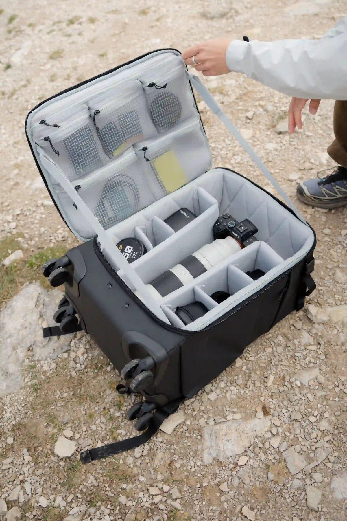 A black camera backpack shown open on a gravel surface, with a person's hand and lower leg visible holding the strap. The bag contains gray interior organizer panels and is filled with camera gear.