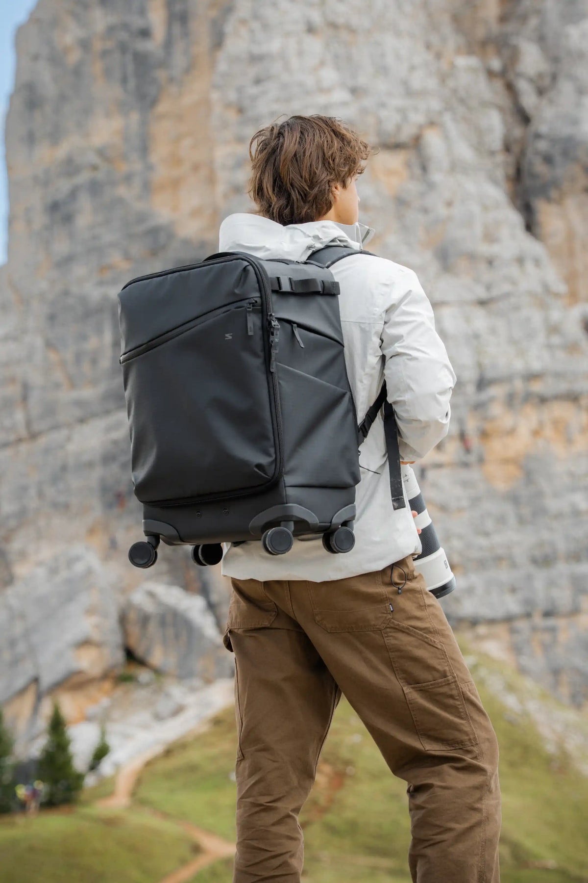 A man seen from behind wearing a large black rolling duffel bag backpack, standing in front of a massive rocky cliffside.