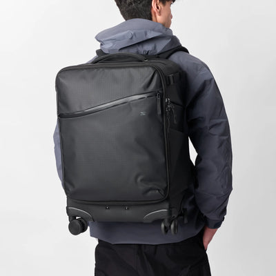 A man wearing a dark gray hooded jacket is shown from the back, carrying a black rolling duffel backpack with multiple compartments and wheels against a light gray studio background.