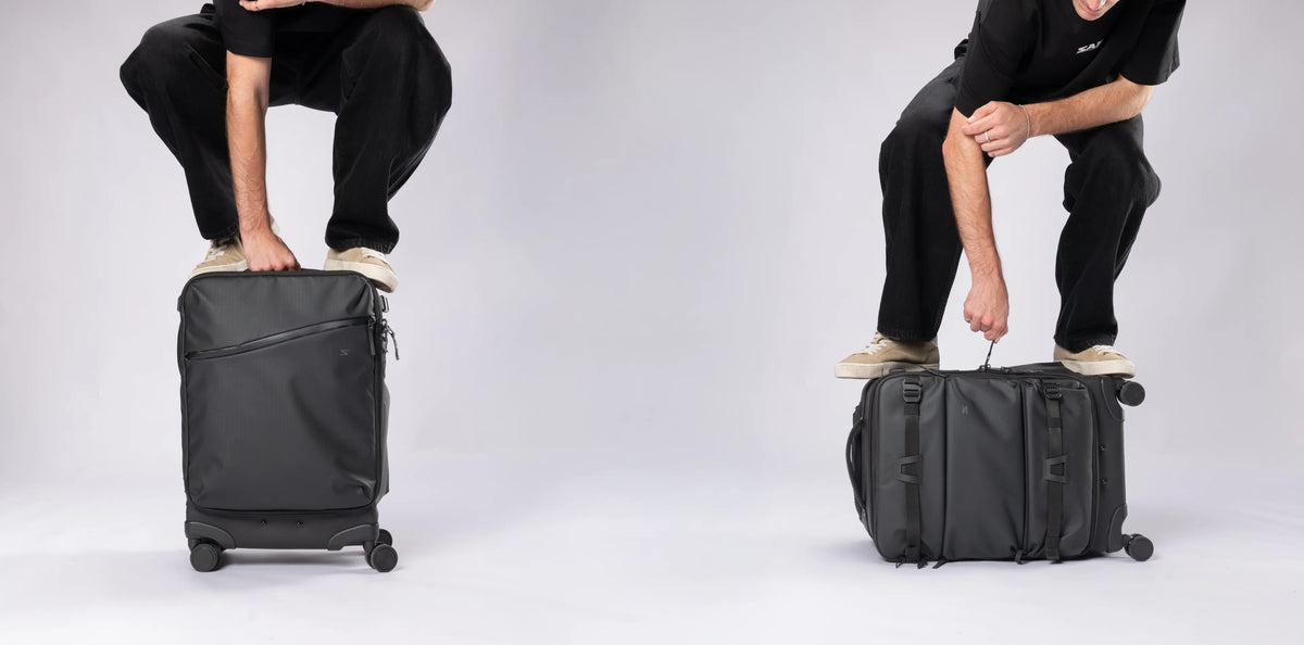 Two views of a black rolling suitcase being pushed by a person in black clothing on a light gray studio background.